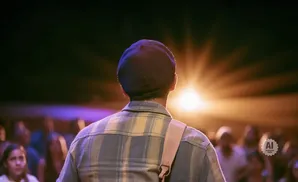 A person in a plaid shirt and hat faces a bright light in front of a blurred audience.