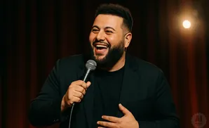 A man with a beard laughs into a microphone, wearing a black suit and t-shirt.