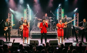 A band performs on stage with singers, guitarists, a bassist, a keyboardist, and a drummer.