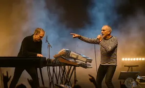 A man in a striped shirt sings into a microphone and points, while another man plays a keyboard on stage with smoke and lights.
