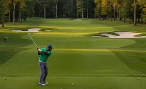 Golfer in green shirt swings at a golf ball on a lush course with trees and sand traps.