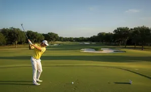 Golfer in yellow shirt swings on a sunny golf course with sand traps and trees.