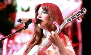 A woman with red hair sings into a sparkly microphone while playing a pink guitar on stage with colorful lights.