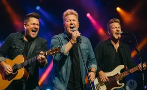Three men on stage with instruments: one plays acoustic guitar, one sings into a microphone, and one plays bass guitar.
