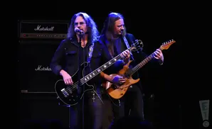 Two guitarists playing on stage. The guitarist on the left plays a black bass guitar and sings into a microphone. The guitarist on the right plays a bro…