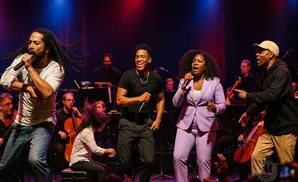 Five singers and an orchestra perform on stage with colorful lighting.
