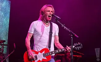 Musician playing a red electric guitar with a white star on a stage with pink lighting.