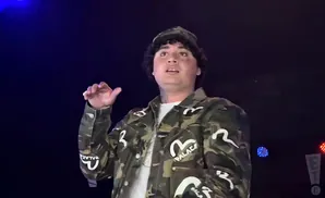 A young man in a camouflage jacket and hat performs on stage under blue lights.