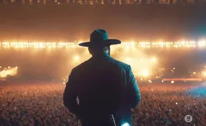 A man in a cowboy hat and jacket stands on a stage facing a large crowd at a concert.