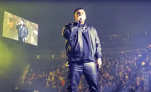 A rapper performs on stage in a black leather jacket and pants, with a large crowd in the background.