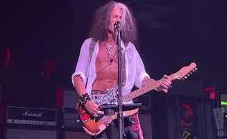 Musician Steven Tyler, shirtless in a white unbuttoned shirt, plays an electric guitar on stage.