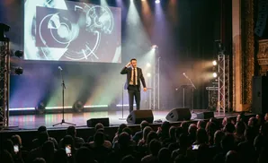 A man in a suit salutes on stage with a screen behind him and an audience watching.