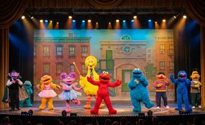 Sesame Street characters perform on stage in front of a backdrop of buildings.