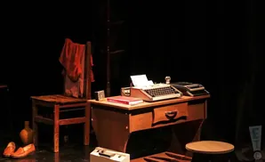 A vintage typewriter on a desk next to a wooden chair with a red garment draped over it.