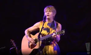 Singer with short blonde hair plays acoustic guitar on stage, wearing a yellow and blue dress.
