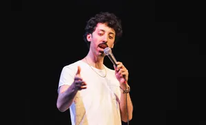 A man with curly hair and a mustache speaks into a microphone on a dark stage.