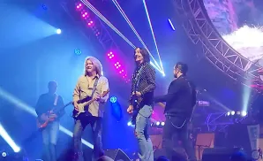 Rock band on stage with blue and pink lights. Two guitarists in front, one in a tan shirt, the other in a polka dot shirt.