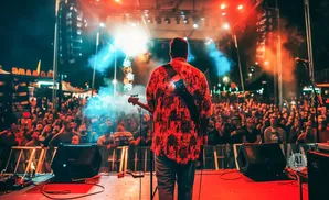 A musician plays bass guitar on a brightly lit stage in front of a large, cheering crowd at night.