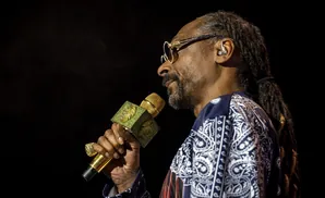 Snoop Dogg on stage holding a gold microphone with diamond accents, wearing glasses and a bandana print shirt.