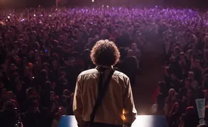 A person with curly hair faces a large, cheering crowd from a stage, with lights illuminating the audience.
