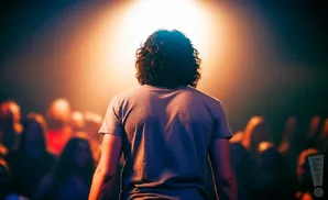 Back view of a person with curly hair under a spotlight, facing a blurred audience.
