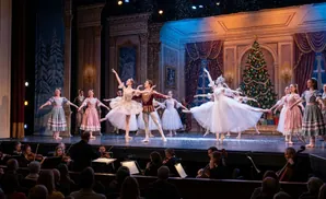 Dancers in elegant costumes perform ballet on a stage adorned with a Christmas tree and falling snow, with an orchestra in the foreground.