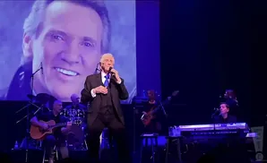 A performer in a suit sings on stage with a projection of his face behind him.