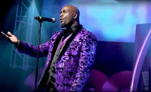 A bald Black man in a sequined purple jacket sings into a microphone on a stage.