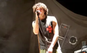 A young man with curly hair sings into a microphone on stage, wearing a sleeveless shirt with a red cross emblem.