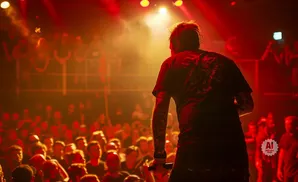A performer with tattoos on their arm stands on a stage, holding a microphone, with a cheering crowd visible in the red-lit background.