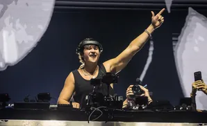 DJ raising a hand in a club with headphones and a necklace.