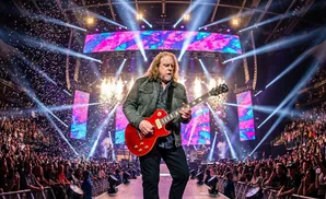 A guitarist plays a red electric guitar on a stage with a large crowd watching and confetti falling.