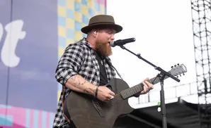 A bearded man in a fedora and checkered shirt plays an acoustic guitar on stage, singing into a microphone.