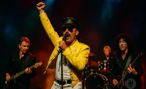 Tribute band member dressed as Freddie Mercury with fist raised, singing into a microphone, flanked by guitarists and a drummer.