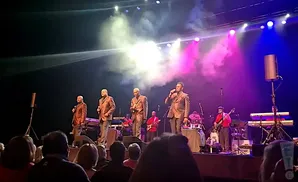 A group of men in patterned suits sing on stage with colorful stage lights and smoke effects.