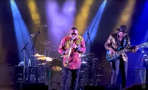 Musicians perform on a stage with colorful lights. A saxophonist in a vibrant shirt stands center, while a bassist in a sequined jacket plays to his right.