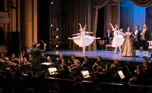 A conductor leads an orchestra as ballerinas and opera singers perform on stage in a grand theater.