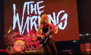 A female guitarist plays a black electric guitar in front of a large screen displaying 