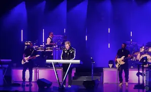 Musicians on stage playing instruments under blue lights, including a keyboardist, guitarist, and drummer.