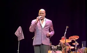 Singer in a purple jacket with a patterned lapel sings into a microphone on stage with a drummer in the background.
