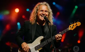 A smiling male bassist with long blonde hair plays a light-colored bass guitar on stage with colorful lights in the background.