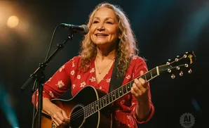A woman with blonde hair smiles while playing an acoustic guitar and singing into a microphone on stage.