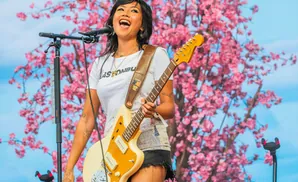 A woman joyfully sings into a microphone while playing a guitar on stage with a pink floral backdrop.