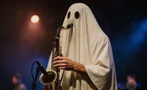 A person in a ghost costume plays a saxophone on stage with a spotlight.