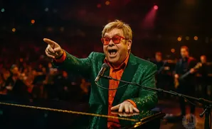 Elton John in a green sequined jacket and orange shirt sings into a microphone while playing a piano.