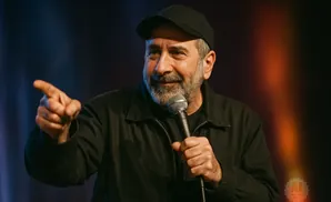A man in a black baseball cap and jacket speaks into a microphone, gesturing with his left hand.