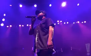 A Black rapper in a black shirt and cap holds a microphone on a purple-lit stage.