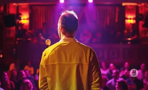 A man in a yellow jacket stands on stage holding a microphone, facing an audience in a dimly lit room.