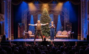 A couple sings onstage at Christmas next to a tree with a snow effect.