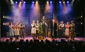 Musicians perform on a rustic stage before a cheering audience, illuminated by stage lights.